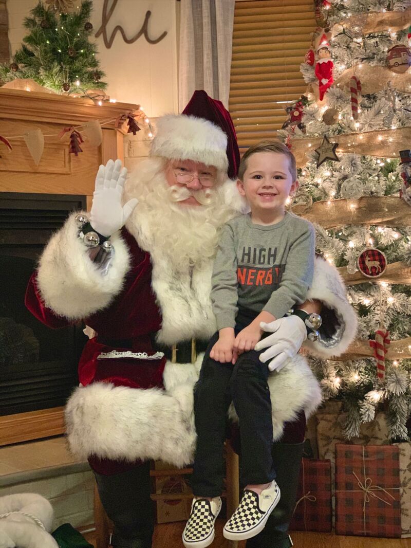 Santa Joe waving beside a Christmas tree with a young boy on his knee during a home visit